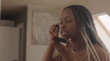Orta ölçekli bir yan çekim. Genç Afrikalı Amerikalı kadın günlük makyajını yapıyor. Kirpiklerine rimel sürüyor ve cep telefonuyla konuşuyor.