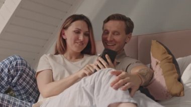 Orta uzunluktaki beyaz kadın erkek arkadaşına yaslanıyor, akıllı telefon kullanıyor, sosyal medyada yayılıyor, gülümsüyor.