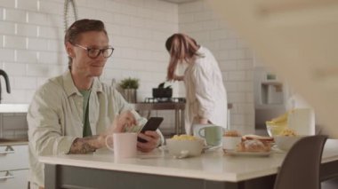 Kafkasyalı bir adam günlük gözlük takıyor, kahvaltı masasında oturuyor, akıllı telefon kullanıyor, karısıyla arka planda yemek pişiriyor.