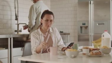 Koyu renk saçlı beyaz bir kadının solunda gözlük takıyor, kahvaltı masasında oturuyor, akıllı telefon kullanıyor, kocasıyla konuşuyor.