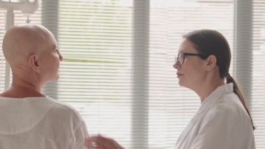 Hastane odasının penceresinin önünde duran kemoterapiden sonra orta yaşlı ve kel bir kadını destekleyen kadın doktorların yavaş çekimleri.