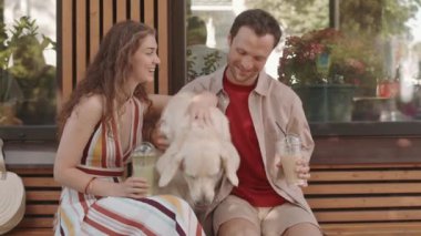 Orta uzunlukta beyaz kadın ve erkek açık havada kahve dükkanında oturmuş, ellerinde buzlu kahve ve matcha tutuyorlar, sevimli köpeklerini okşuyorlar.
