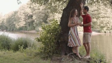 Çıplak ayaklı beyaz kadın ve yaz kıyafetleri giyen bir adam göletin yanında durup bardaktan beyaz şarap içerken, sohbet ederken geniş bir kare.