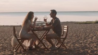 Kafkasyalı mutlu kadın ve erkek kumlu deniz kıyısında oturmuş, beyaz şarapla romantik bir akşam yemeği yiyorlar.