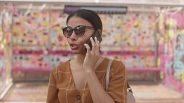 Modern melez bir kadının göğüs göğsü. Yazlık giysiler ve güneş gözlüğü takıyor, cep telefonuyla konuşuyor, gülümsüyor.
