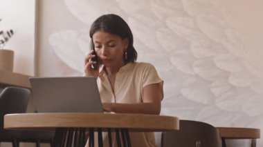 Restoranda masada oturan, cep telefonuyla konuşan, taşınabilir bilgisayar kullanan meşgul genç bir kadının orta boy yakın çekimi.