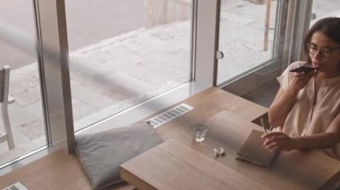 Kafede masada oturan, cep telefonu hoparlöründen konuşan, melez, zeki genç bir kadının yüksek açılı görüntüsü.