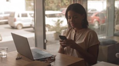 Kafede dizüstü bilgisayarlı masada oturan, akıllı telefondan mesaj atan ve gülümseyen melez genç bir kadının orta boy yakın çekimi.