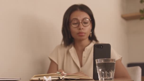 低角度的混血女商人戴着眼镜坐在桌旁 视频在手机上 设备靠在玻璃杯上 — 图库视频影像