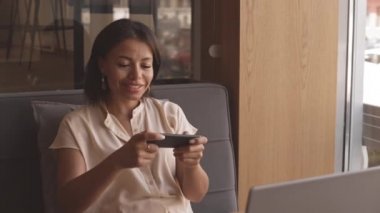 Kafede oturan, akıllı telefondan video oyunu oynayan, gülümseyen neşeli melez kadının orta boy yakın çekimi.