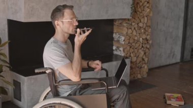 Tekerlekli sandalyede oturan, dizüstü bilgisayar ve akıllı telefon kullanarak çalışan beyaz bir erkek.