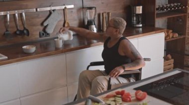 Mutfağında tekerlekli sandalyede, bardağa süt döken ve içen melez genç bir kadın.