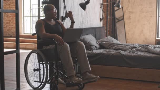 ベッドルームの車椅子に座って膝の上にノートパソコンを持ちスピーカーフォンで話している短髪の女性の完全なショット — ストック動画