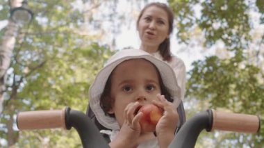 Low-angle shot of cute toddler boy with curly brown hair eating apple while sitting in stroller outdoors on sunny day. His mother in blurred background smiling while talking to him