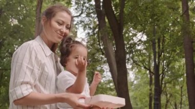 Low-angle medium shot of happy mom and toddler son enjoying summer outdoors in park, having fun eating sweet cherries