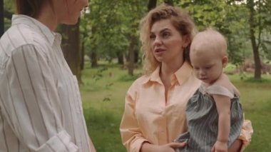 Medium shot of young woman with pretty little daughter in hands talking to female friend standing outdoors in park on summer day