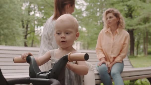 可爱的白种人幼儿小女孩在公园玩三轮车时 妈妈在长椅上与女友进行背景对话 — 图库视频影像