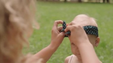 Medium close-up of woman tying polka dot bow over head of her cute toddler daughter spending time outdoors in park in summer