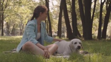 Full shot of happy Caucasian woman wearing light-blue blazer, sitting on lawn in sunny park, petting her cute grown-up dog and talking to pet