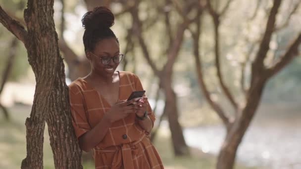 Медиум Запирал Счастливую Африканскую Женщину Стоящую Опираясь Дерево Солнечном Парке — стоковое видео