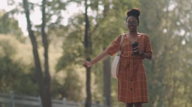 Medium long of beautiful modern African woman holding smartphone in hand, standing on side of road, calling cab with thumbs up gesture in city near park