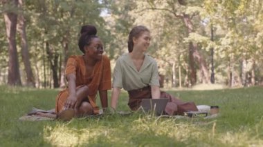 Wide shot of African and Caucasian female friends sitting on lawn in park, using laptop computer, looking at dog running around, smiling and talking