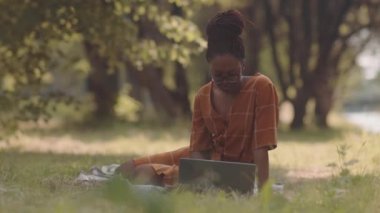 Full shot of young African woman wearing summer dress, using portable computer, sitting on grass in park
