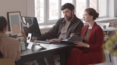 Orta uzunlukta beyaz kadın ve erkek iş arkadaşları gündüz masa başında otururken, adam bilgisayar ve kadın kullanarak not defteri tutuyor.