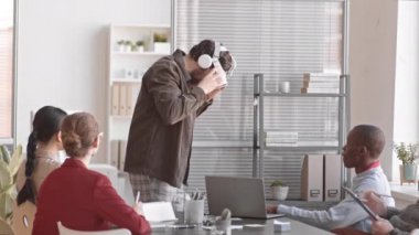 Orta boylu, sakallı, kafkasyalı, VR kulaklık takmış, çeşitli meslektaşları konferans masasında onu izliyor.