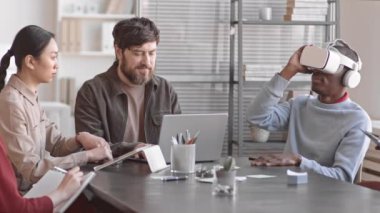 BT şirketinin ofisindeki konferans masasında oturan, iş arkadaşlarıyla tecrübelerini paylaşan, VR kulaklık takan Afrikalı genç bir adam.