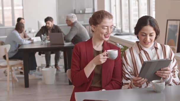 白种人女人穿着红色的正装 喝着热茶 和她的女混血儿朋友用平板电脑聊天 坐在办公室的办公桌前 — 图库视频影像