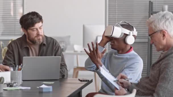 Waist-up of young African man wearing VR headset sitting at conference table in office of IT company, sharing experience with coworkers who taking notes