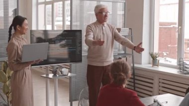 Orta uzunluktaki duygusal kıdemli beyaz erkek ve genç Asyalı kadının bilgisayar koduyla ekranın kenarlarında durarak konferans masasında oturan meslektaşlarına sunum yapmaları.