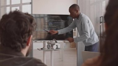 Orta uzunluktaki heyecanlı Afrikalı bilişim uzmanı sunum yapıyor. Konferans odasında oturan tanınmayan yatırımcılara ekranda bilgisayar kodlarını gösteriyor.