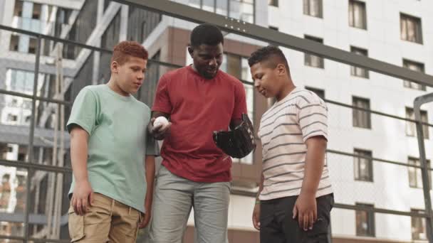 中低角镜头 年轻的非洲裔美国人戴着棒球手套 向他的两个儿子解释如何接球 听他说话 一起站在室外运动场上 — 图库视频影像