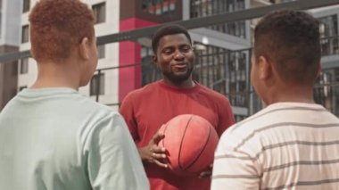 Güneşli bir günde açık havada Afrika kökenli Amerikan basketbol koçunun antrenmanlarını dinleyen iki gencin dikiz görüntüsü.