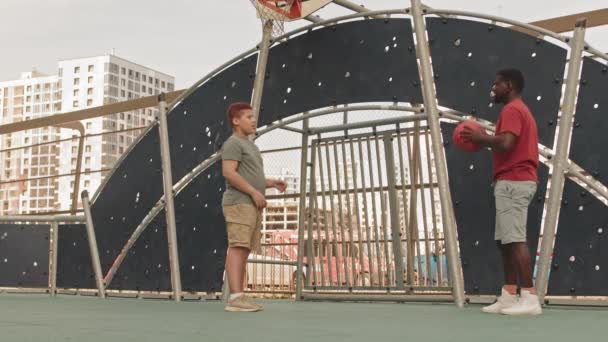 アフリカ系アメリカ人の男と彼の十代の息子の広いショットは スポーツグラウンド投げバスケットボールでお互いに屋外に立って 暖かい晴れた日に話して — ストック動画