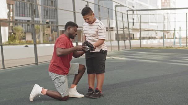 一名身穿运动服的年轻的非洲裔美国男子在训练前帮助他十几岁的儿子戴棒球手套 夏日他站在运动场外 — 图库视频影像