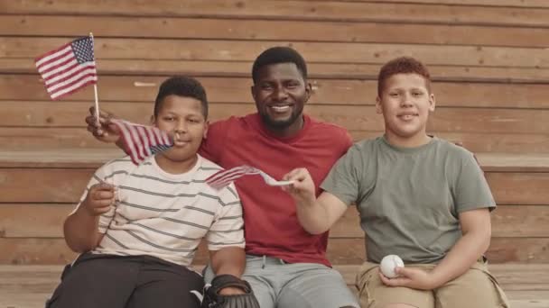 在阳光灿烂的日子里 一个快乐的非裔美国男人和他的两个年幼的儿子坐在运动场外 对着摄像机微笑 挥动着美国国旗 — 图库视频影像