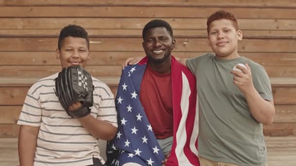アフリカ系アメリカ人の男が肩にアメリカ国旗を掲げてカメラに向かって笑っている姿を 野球と手袋をしてポーズをとっているふたりの10代の息子が — ストック動画