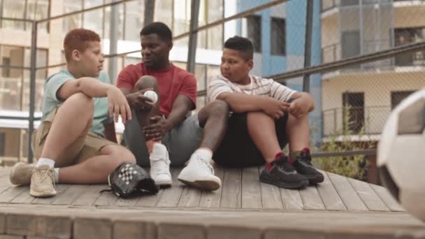 若いですアフリカ系アメリカ人男性と2混合レースティーン少年たち座っています上のフェンスで囲まれたスポーツグラウンドチャット上の晴れた日 — ストック動画