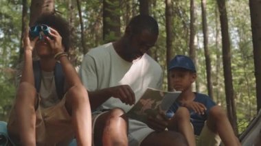 Ormanda kamp yapan iki küçük oğlu olan Afrikalı Amerikalı bir adamın orta boy fotoğrafı. Bir çocuk dürbünle bakarken diğeri haritayı inceliyor ve babası ağaçların altında oturuyor.