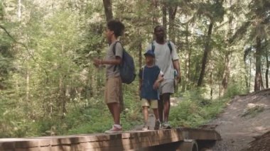 Omuzlarında sırt çantaları olan iki Afrikalı Amerikalı çocuk ve babalarının, yaz ormanı boyunca ahşap köprü boyunca yürüyüp yol haritasına bakarken tam boy çekimleri.