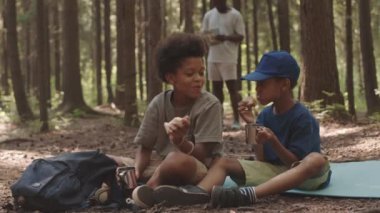 Babaları arka planda harita incelerken iki Afro-Amerikan kardeşin kamp alanında çayla ve sandviçle yılan gibi kıvrılıp oturmasının yavaş çekimi.