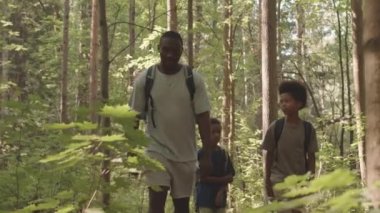 Afrika kökenli Amerikalı bir adam ve sırt çantalı iki küçük oğlunun kamp gezisi sırasında ormanda yürürken konuştuklarını gösteren yavaş çekim.