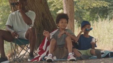 İki neşeli Afrikalı Amerikalı çocuğun ve babalarının ormanda kamp yaparken, büyük bir ağacın altında oturup, sohbet ederken ve dürbünle bakarken orta boy fotoğrafları.
