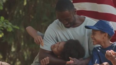 Afrika kökenli Amerikalı babayı ve küçük tatlı oğullarının büyük bir ağacın altında kucaklaşıp dinlenirken, Amerikan bayrağının asılı olduğu orta boy bir fotoğraf.