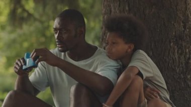 Kamp gezisi sırasında ağaçların altında birlikte oturan kuşları ve hayvanları izlemek için dürbün kullanan Afrikalı Amerikalı bir adamın orta boy fotoğrafı.