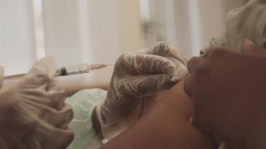 Koluna SARS-CoV-2 aşısı yaptıran Afro-Amerikalı kadının modern doktor ofisinde otururken çekilmiş el kamerası görüntüsü.