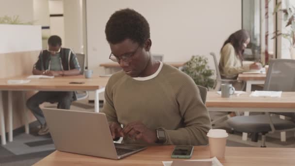 在同时代的办公室里 坐在办公桌前的年轻黑人聚精会神地在笔记本电脑上打字 而他的同事则背景地做文书工作 — 图库视频影像